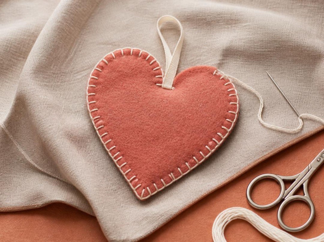 Overhead photograph of a flat terracotta-rose felt heart with ivory blanket stitching around most of its curved perimeter, a cotton hanging loop, and a small unstitched opening for stuffing.